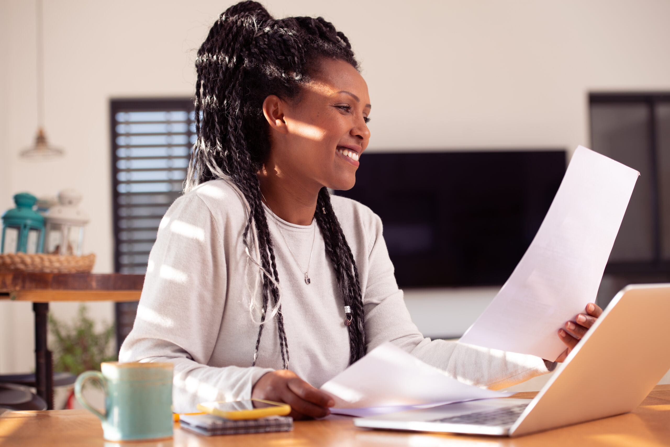 African-American woman sitting at the desk with laptop, reading, reviewing few documents. Female working from distance at the home office. Online business career.