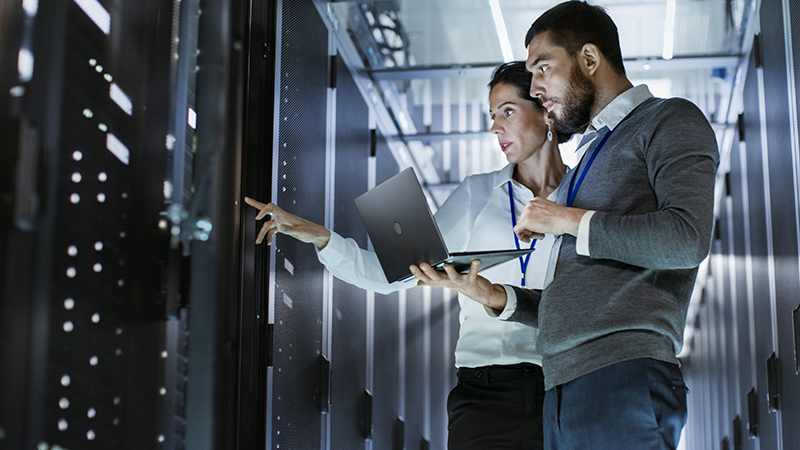 Male IT Specialist Holds Laptop And Discusses Work With Female Server Technician. They’re Standing In Data Center, Rack Server Cabinet Is Open.